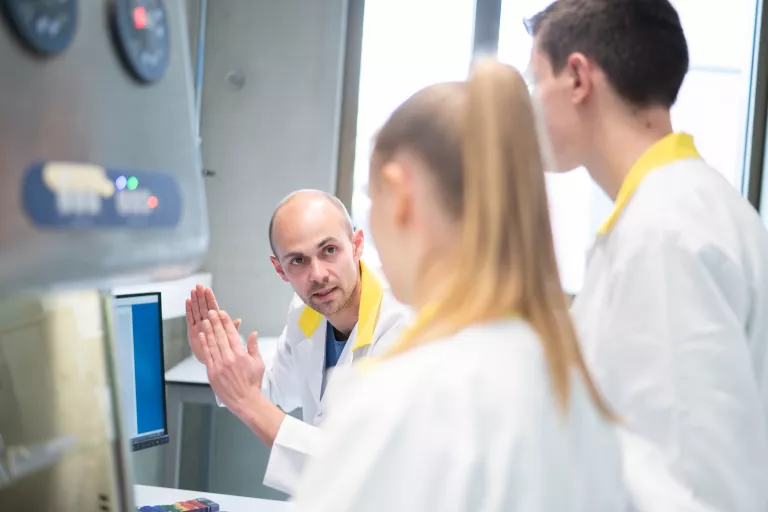 Someone explaining a piece of lab technology to two other researchers, all wearing lab coats